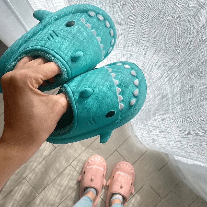 A playful pair of Sharkmo shark slippers in vibrant teal, featuring a whimsical shark face design, gripped by a hand against a soft-hued backdrop – find your bite of fun footwear fashion at sharkmo.shop.